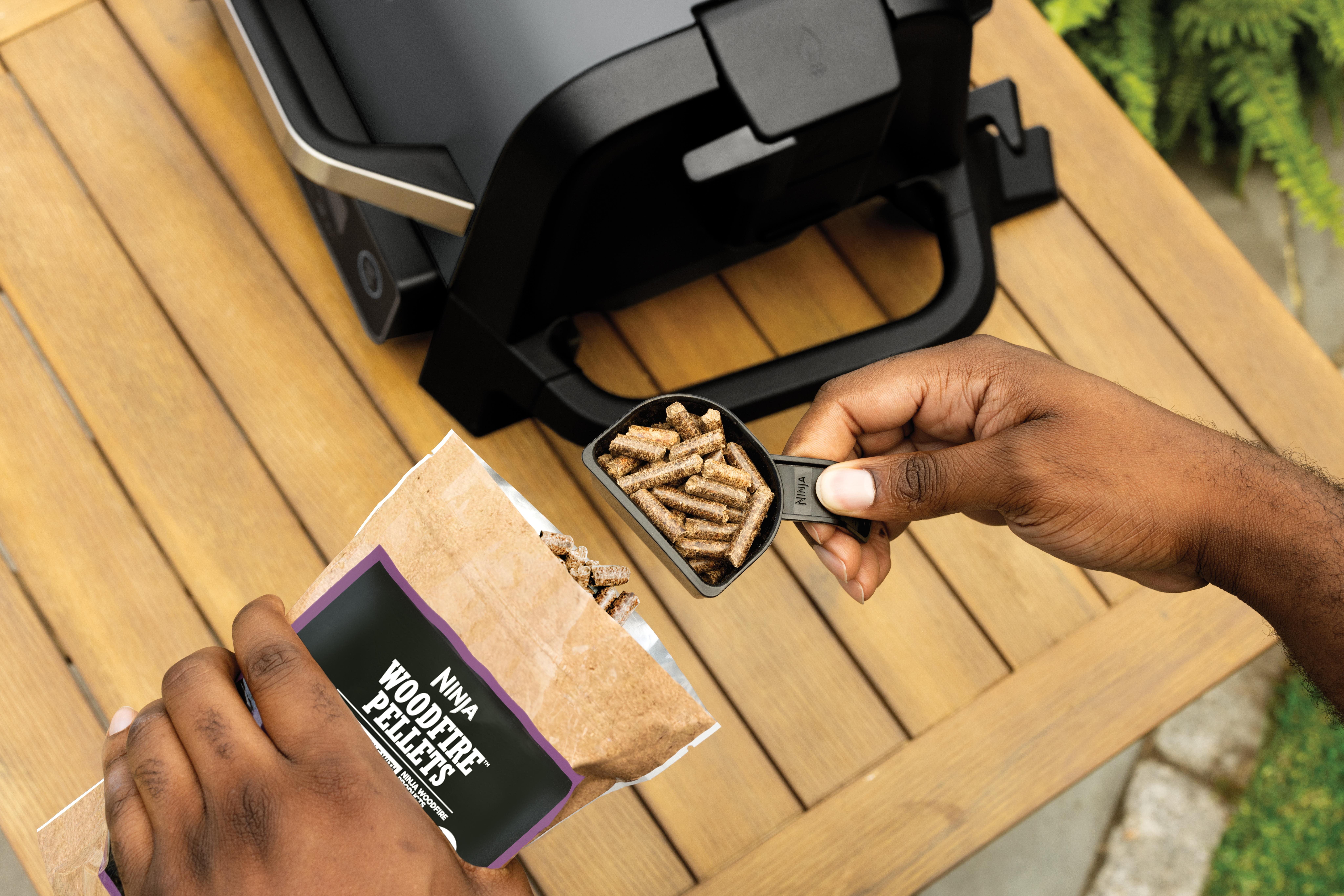 Eine Person gießt Woodfire-Pellets aus einem Beutel in einen schwarzen Messlöffel, über einem Holztisch. Ein Ninja-Grill ist sichtbar.