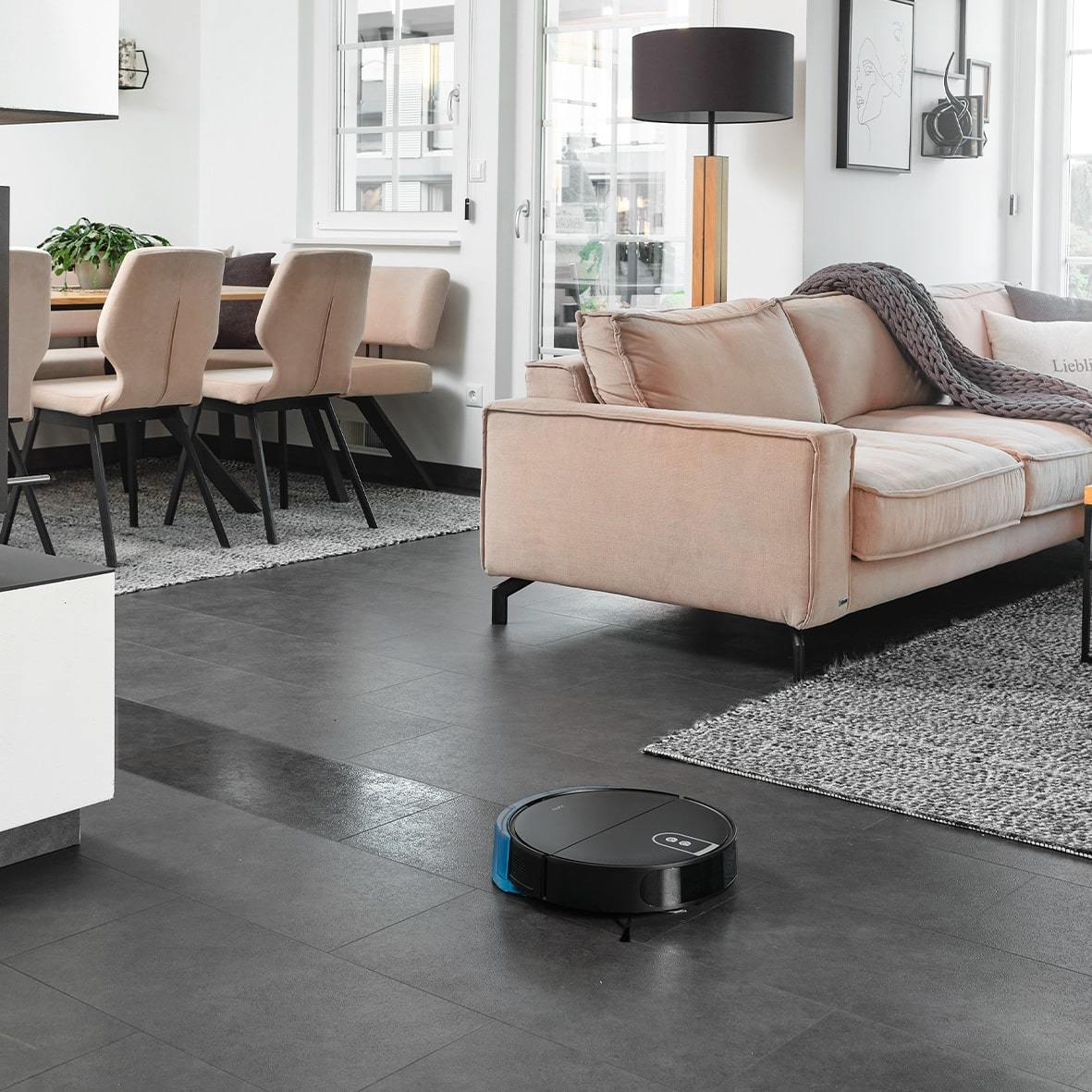 Un aspirapolvere robotico nero su un pavimento scuro in un soggiorno. C'è un divano e un tavolo.
