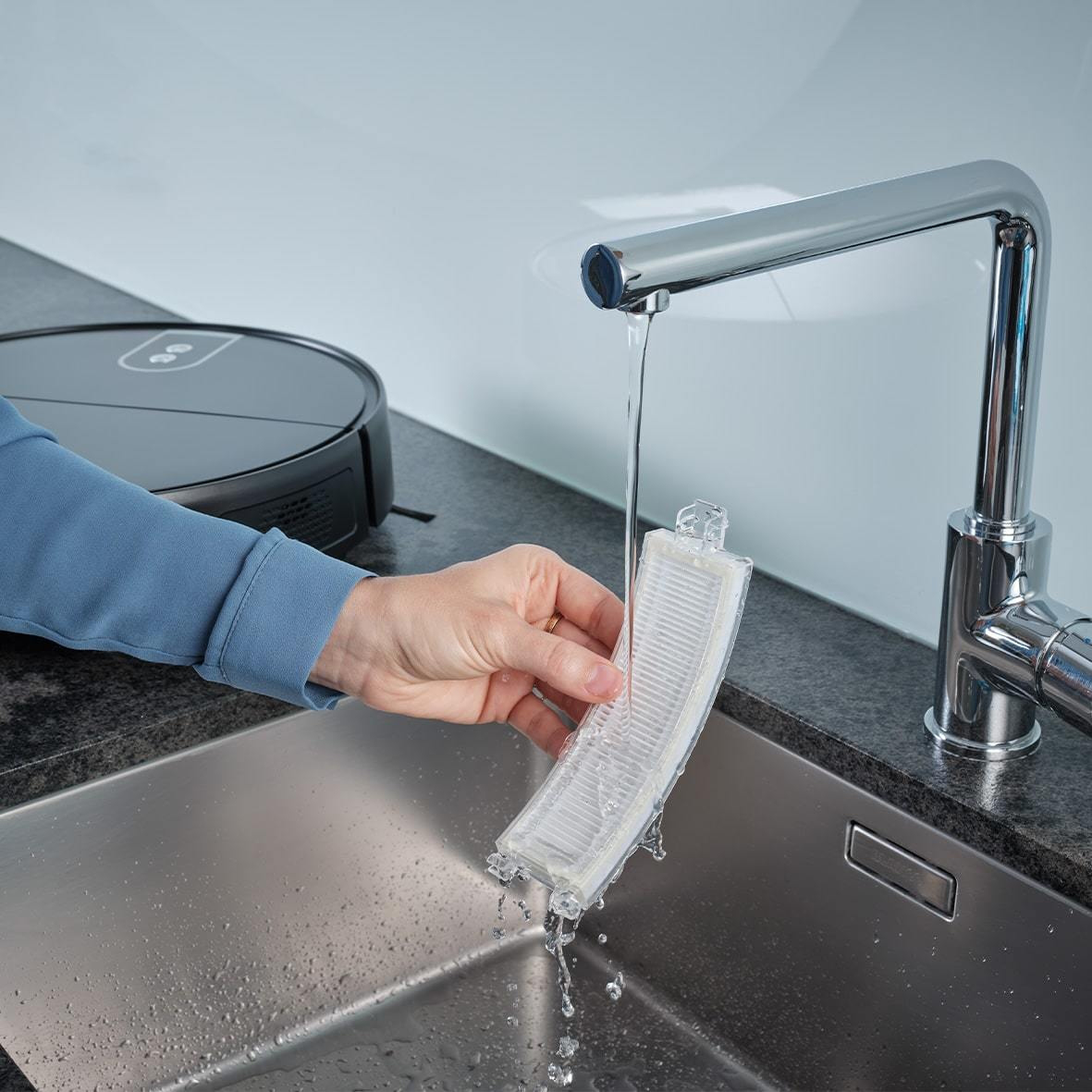 Una mano tiene un filtro sotto un rubinetto aperto, un aspirapolvere robot nero nelle vicinanze.