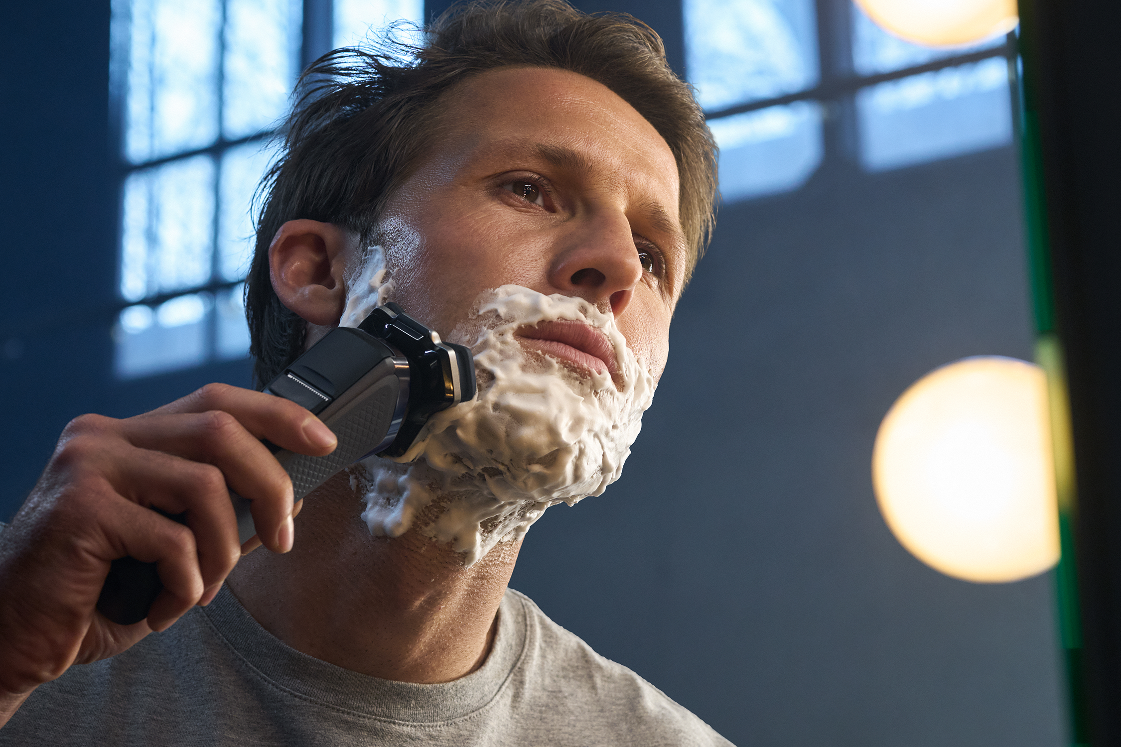 Man shaves with electric razor, foam on face. Background: mirror, window.