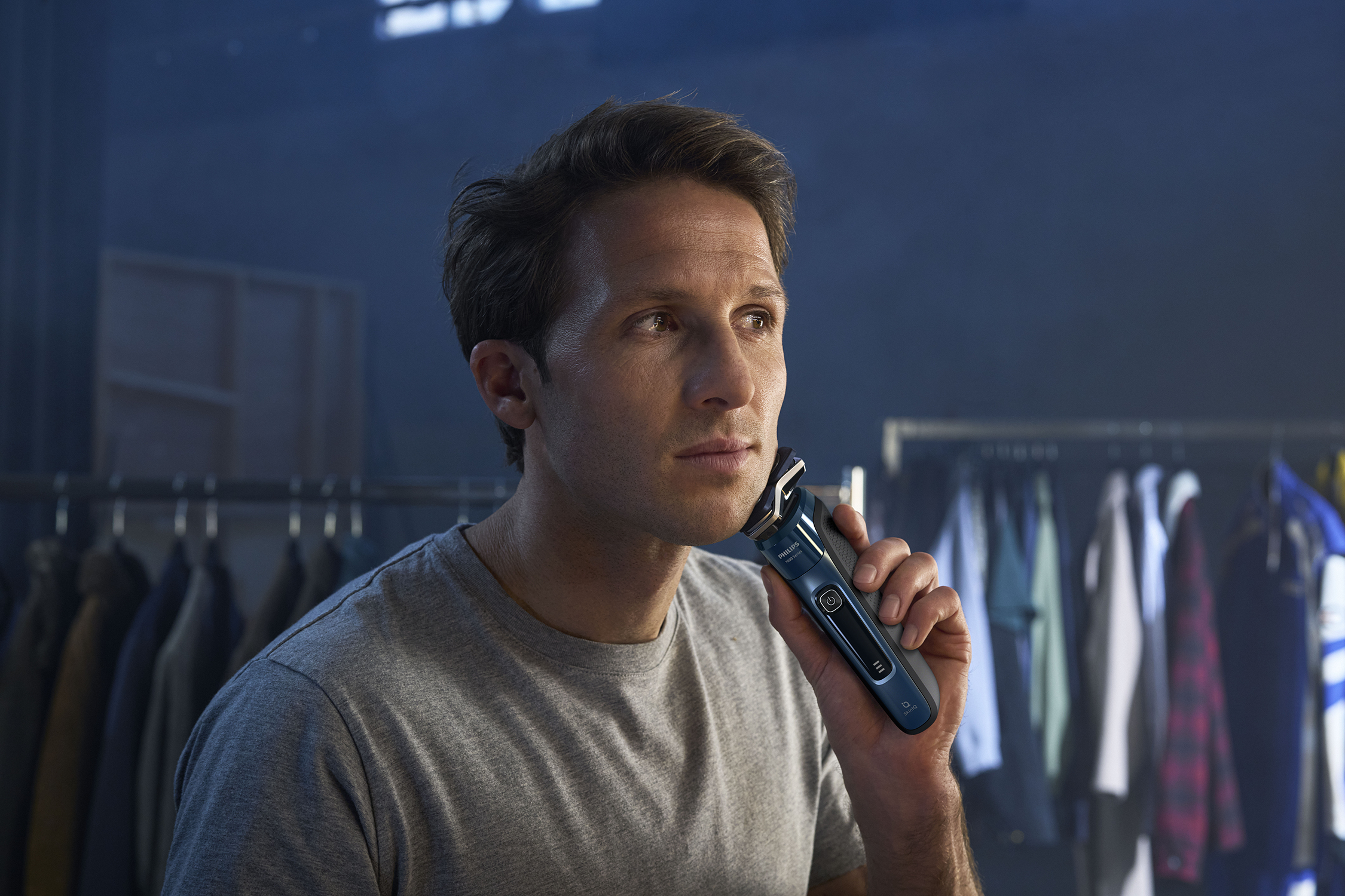 Man shaving his face with a blue and black electric shaver. He is in a room with clothes.
