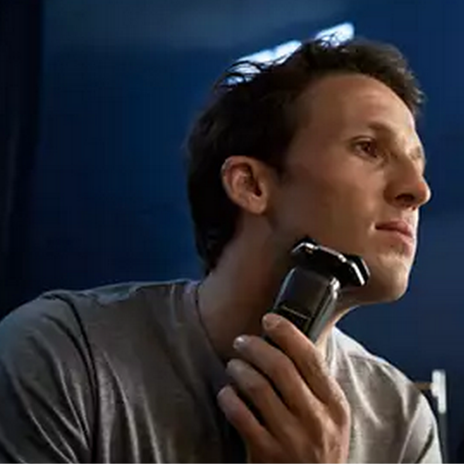 A man shaves his face with an electric razor. Dark background.