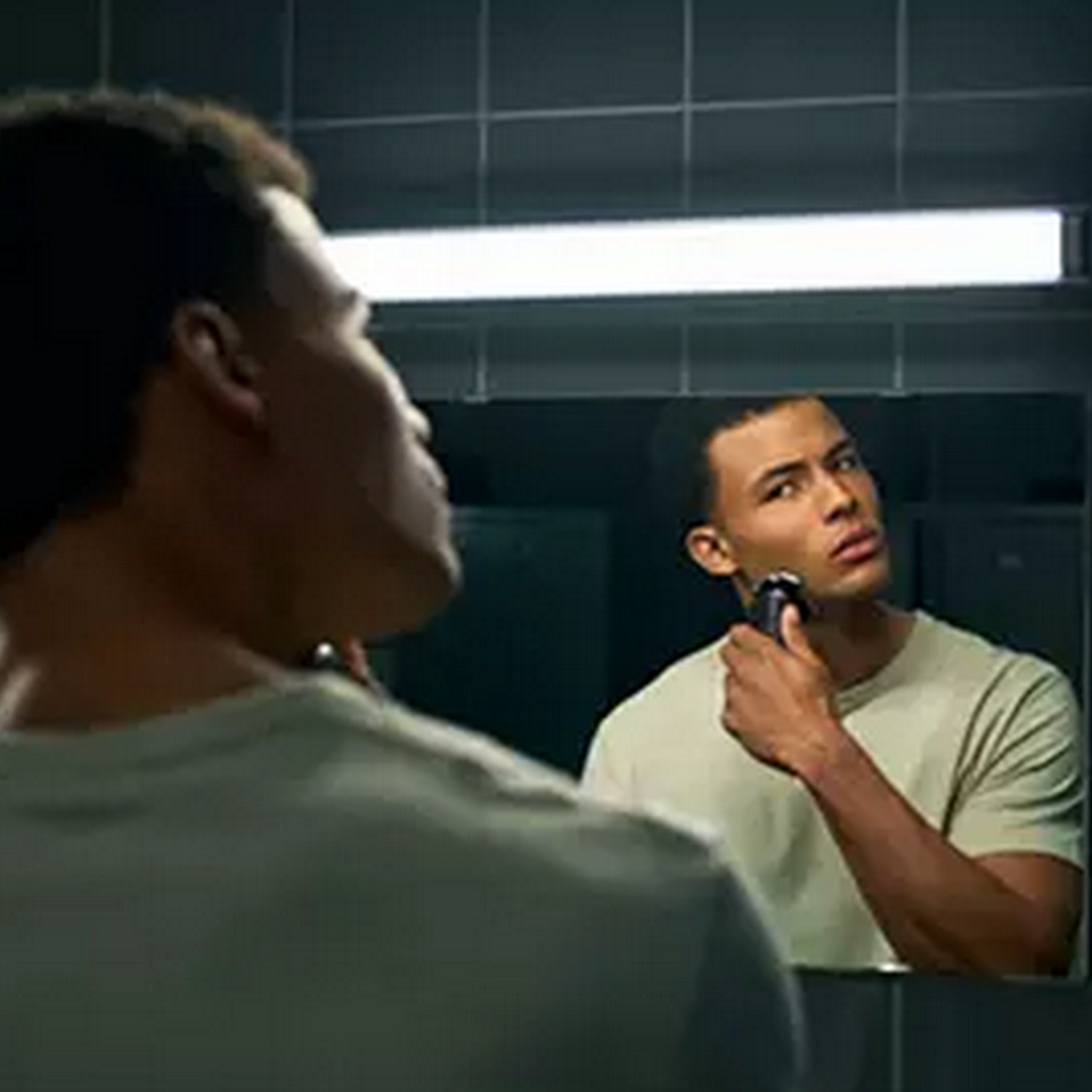 A man shaves his face in a mirror. He is looking at his reflection. The background is a bathroom.