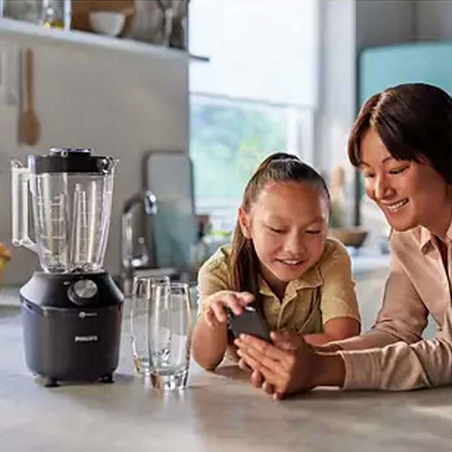 A woman and a girl look at a phone. A blender and glasses are on the counter. Kitchen background.