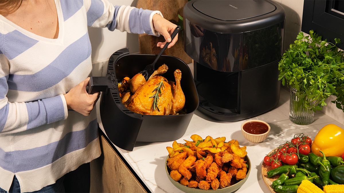 Mujer usando freidora de aire con pollo cocido y guarniciones en una encimera de cocina.
