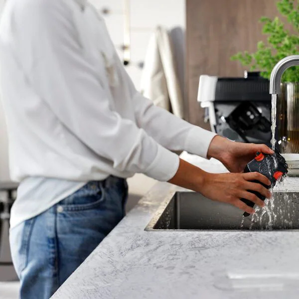 A person rinses a black and orange object under running water in a kitchen sink.