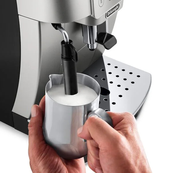 A person pours milk into a stainless steel pitcher from a coffee machine. The machine is silver and black.