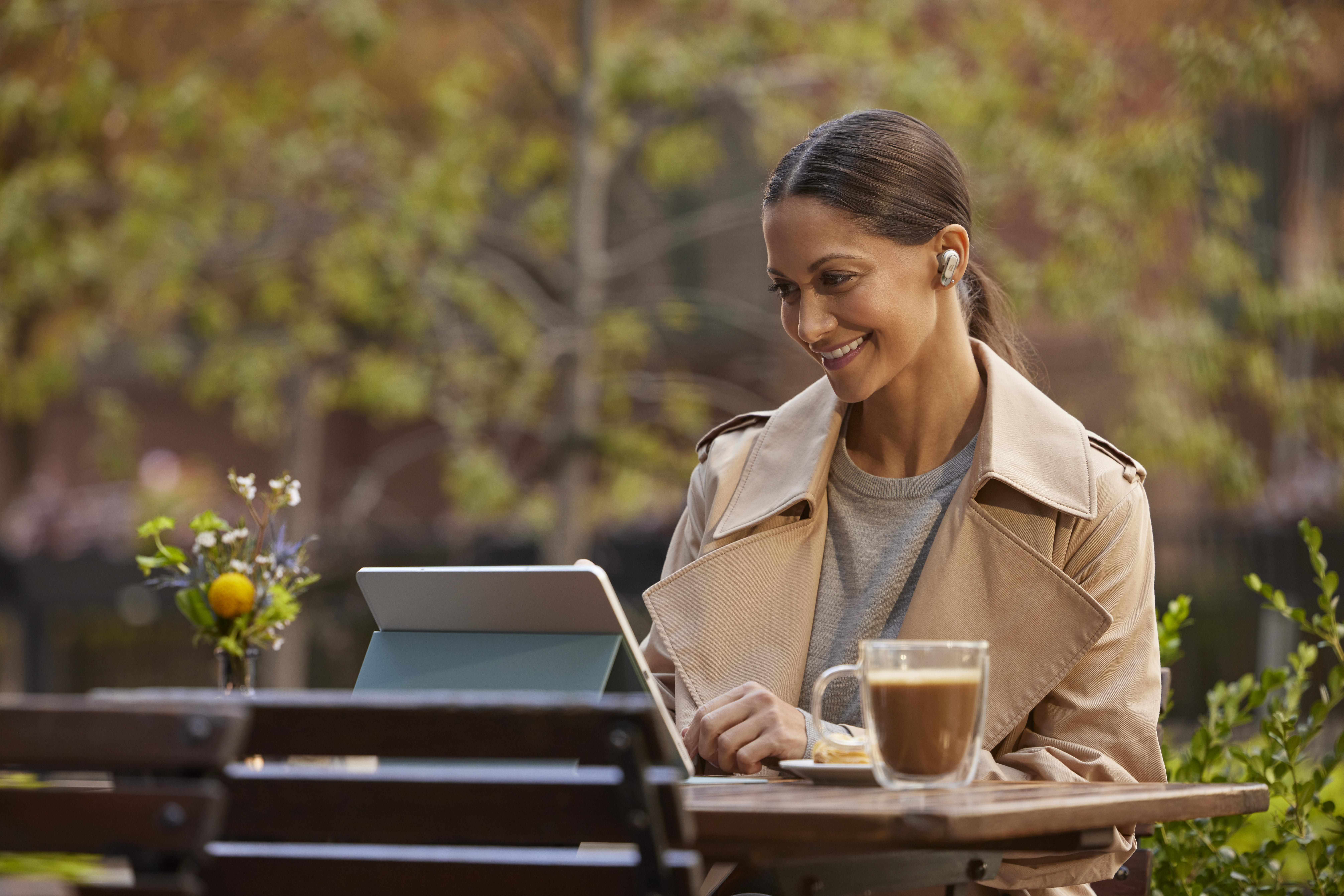 Eine Frau sitzt im Freien, benutzt ein Tablet und Ohrhörer, lächelt, mit Kaffee und Blumen.