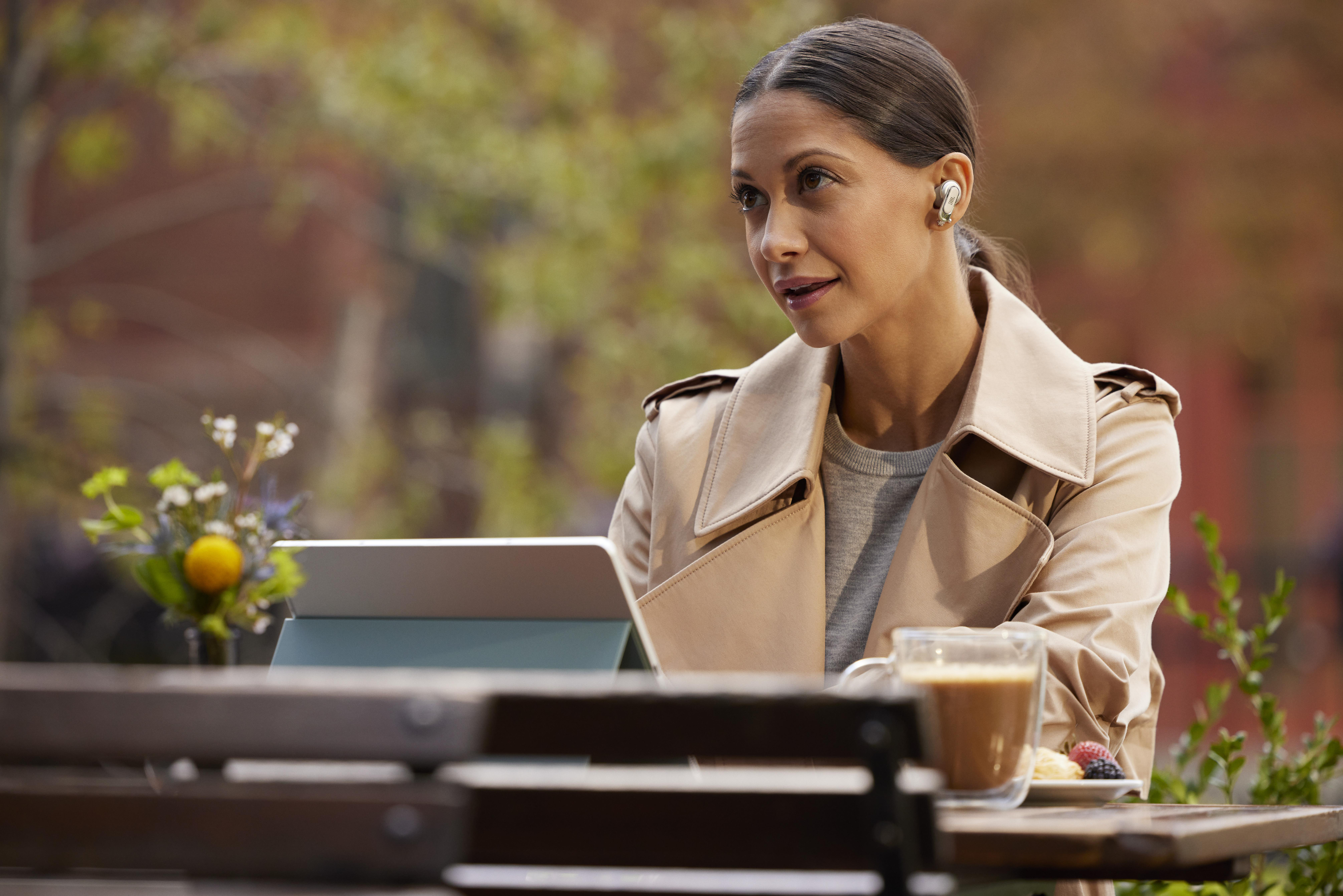 Frau in einem Café, mit Kopfhörern und Mantel, schaut auf ein Tablet. Kaffee und Gebäck auf dem Tisch.