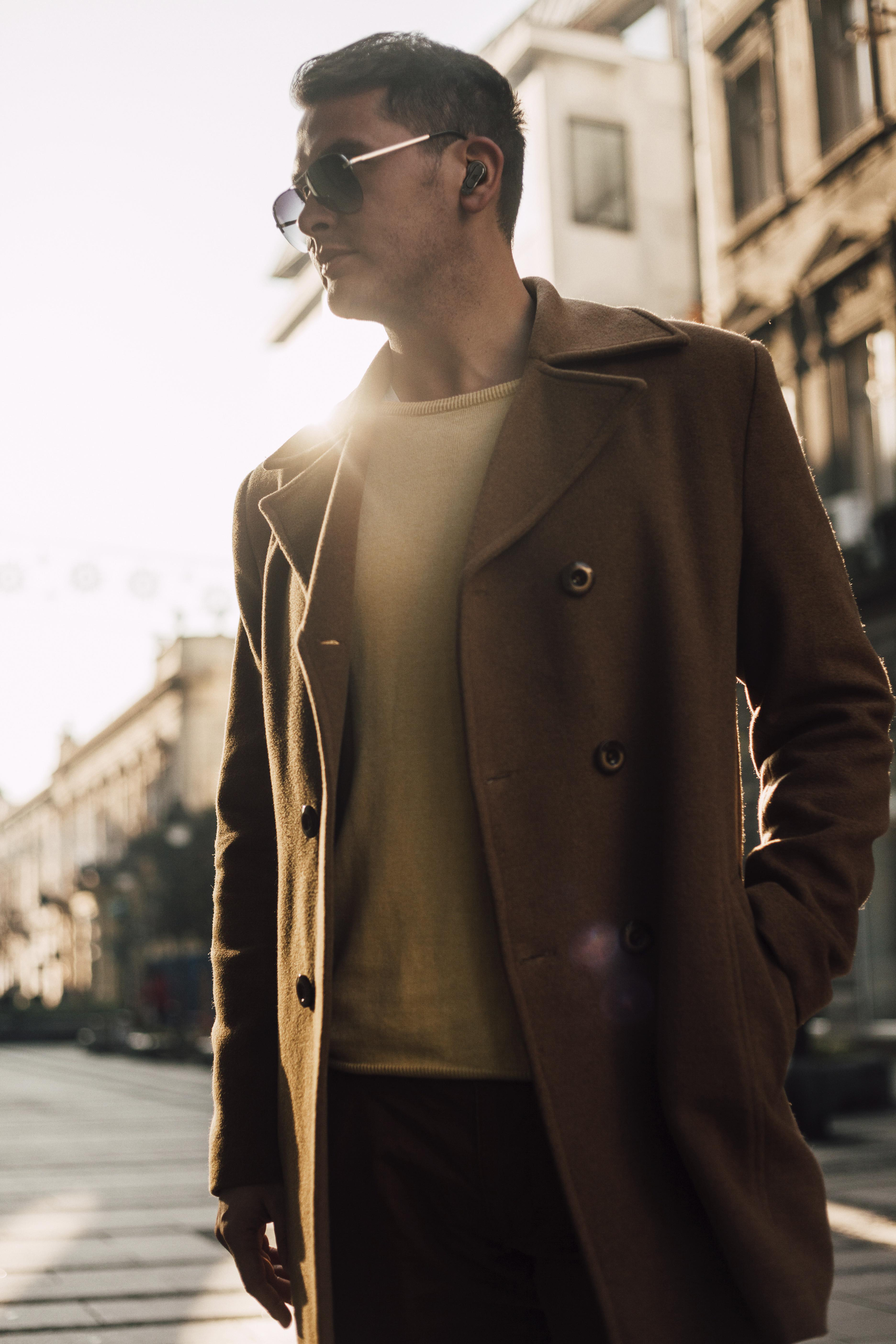 Mann mit Sonnenbrille und braunem Mantel, der sich im Freien bewegt. Die Sonne scheint.