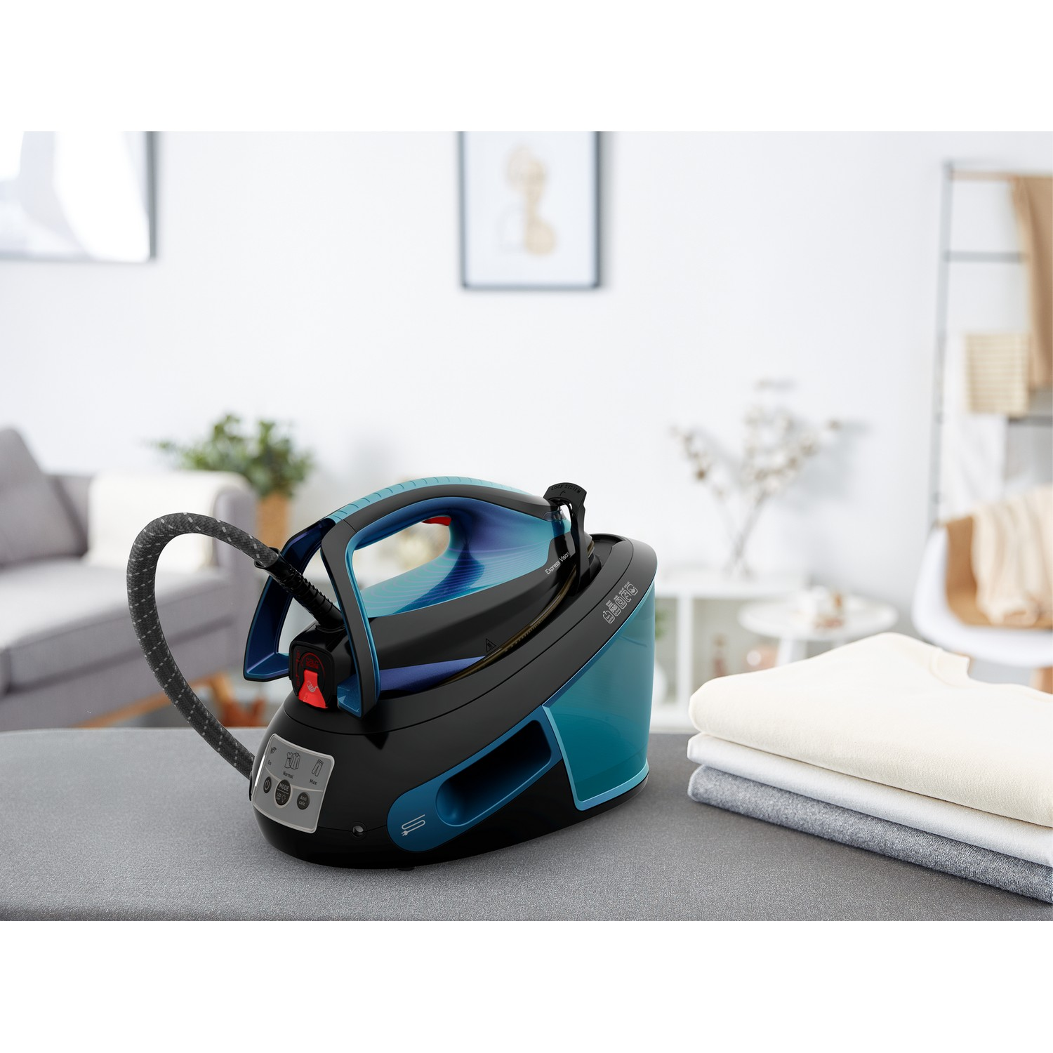 Black and teal iron station on a gray surface with folded clothes on the right. A white wall is in the background.