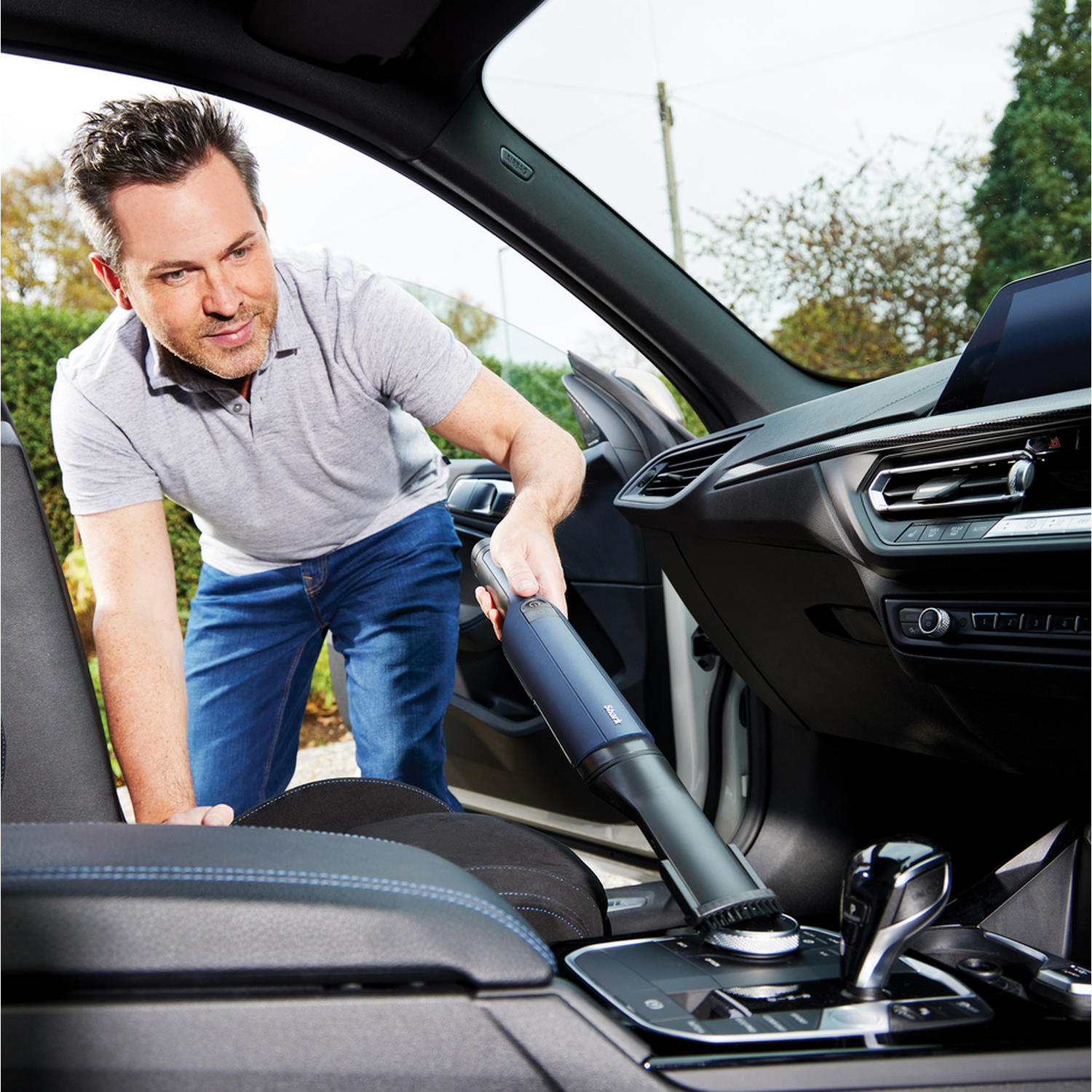 Man cleaning the inside of a car with a handheld vacuum cleaner near the gear shift and dashboard.