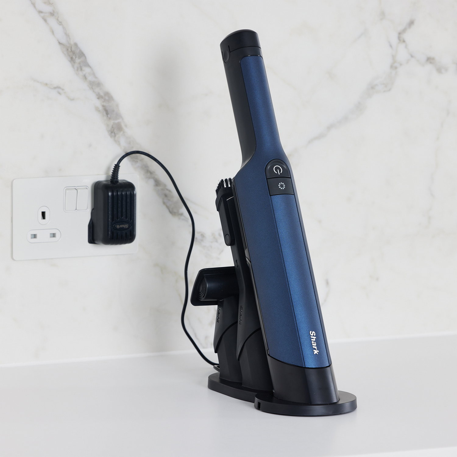 A blue and black cordless vacuum cleaner with a charging station. It's on a white surface near a socket.
