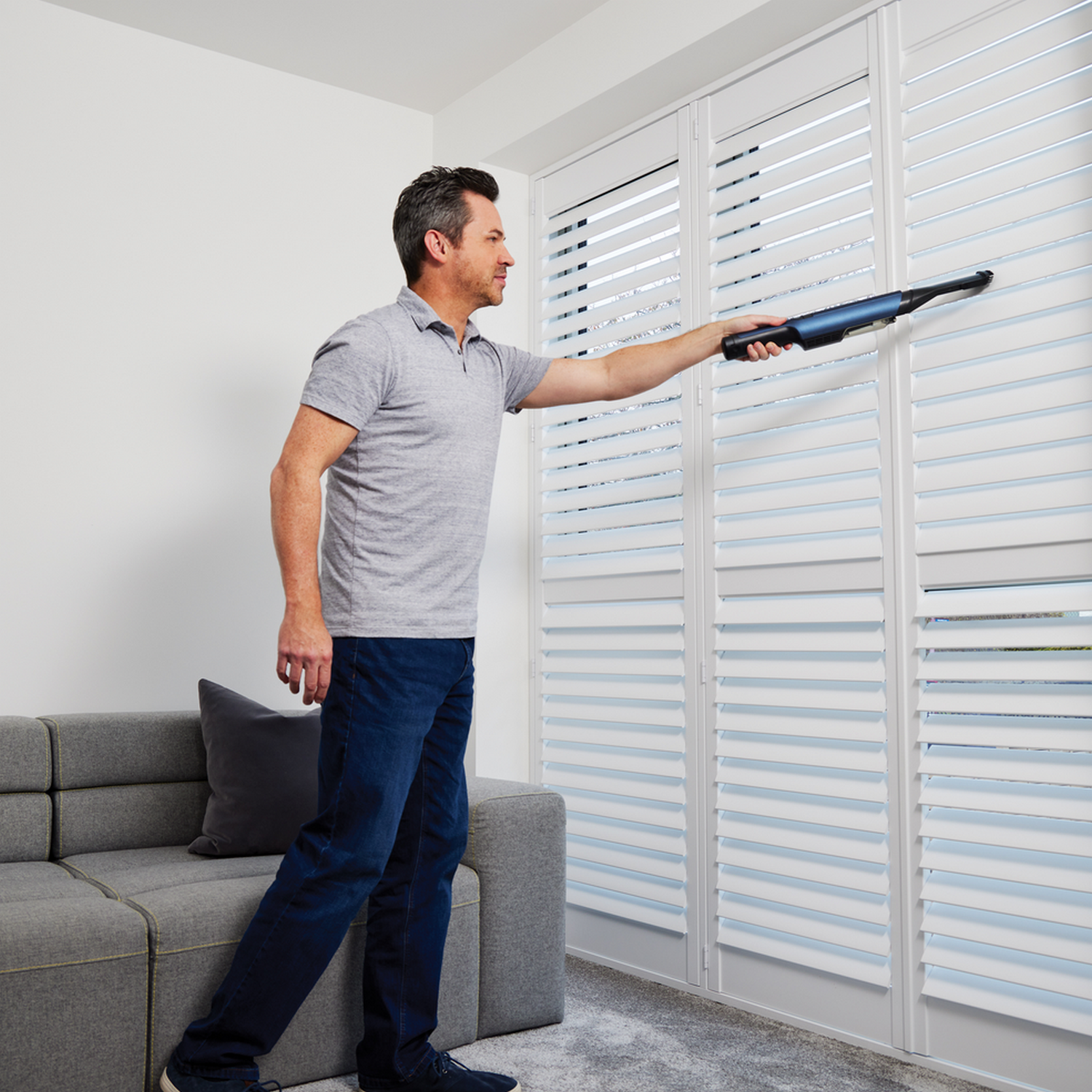 A man cleaning white shutters with a handheld vacuum cleaner.