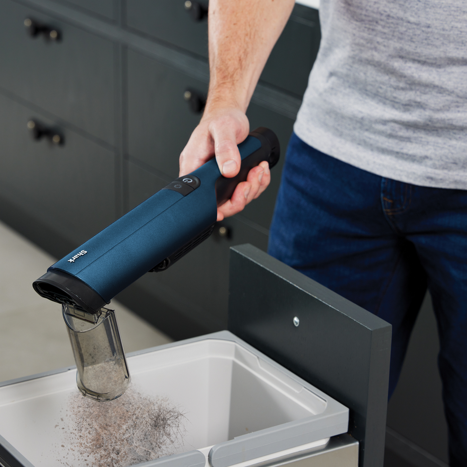 A person emptying a blue handheld vacuum cleaner into a trash can with a grey lid.