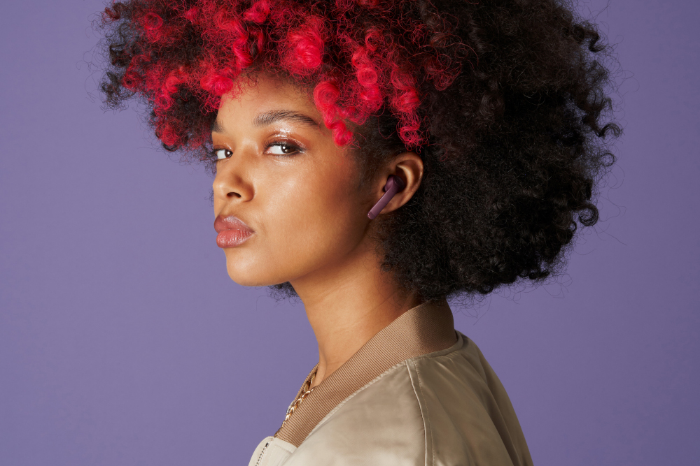 Femme aux cheveux bouclés rouges et noirs porte des écouteurs. Elle regarde par-dessus son épaule sur fond violet.