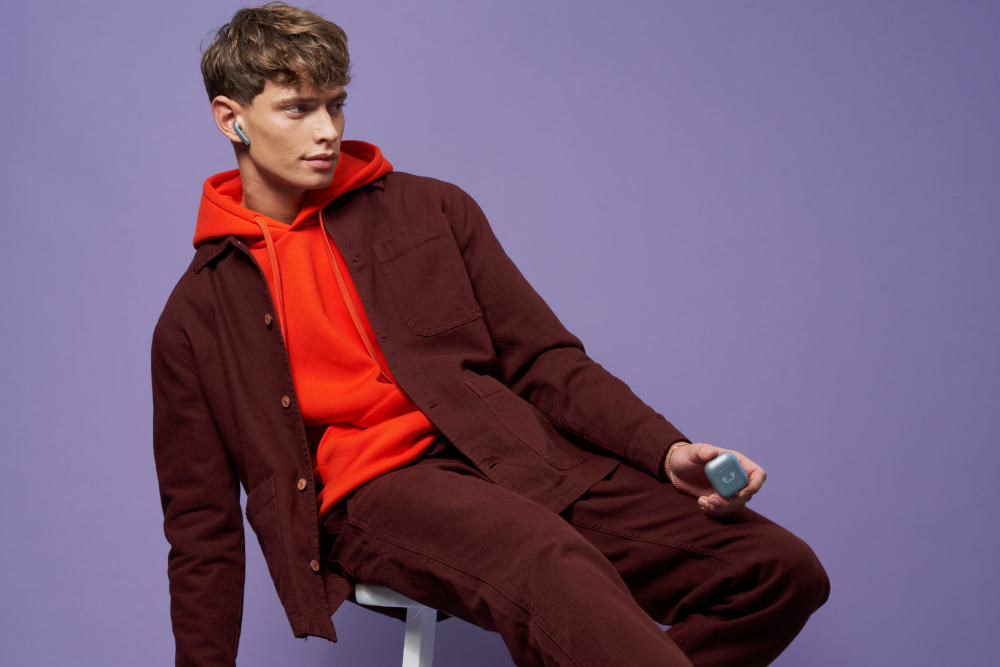 Un homme en sweat à capuche rouge et veste marron tenant un écouteur bleu, posant sur fond violet.