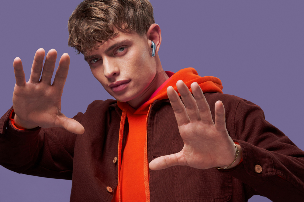 Un jeune homme portant des écouteurs et un sweat à capuche orange fait des gestes avec ses mains.