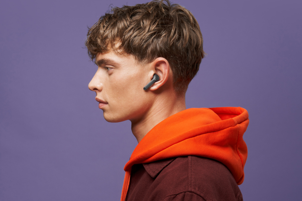 Een jonge man met een oranje hoodie draagt een grijze draadloze oordop in zijn rechteroor.