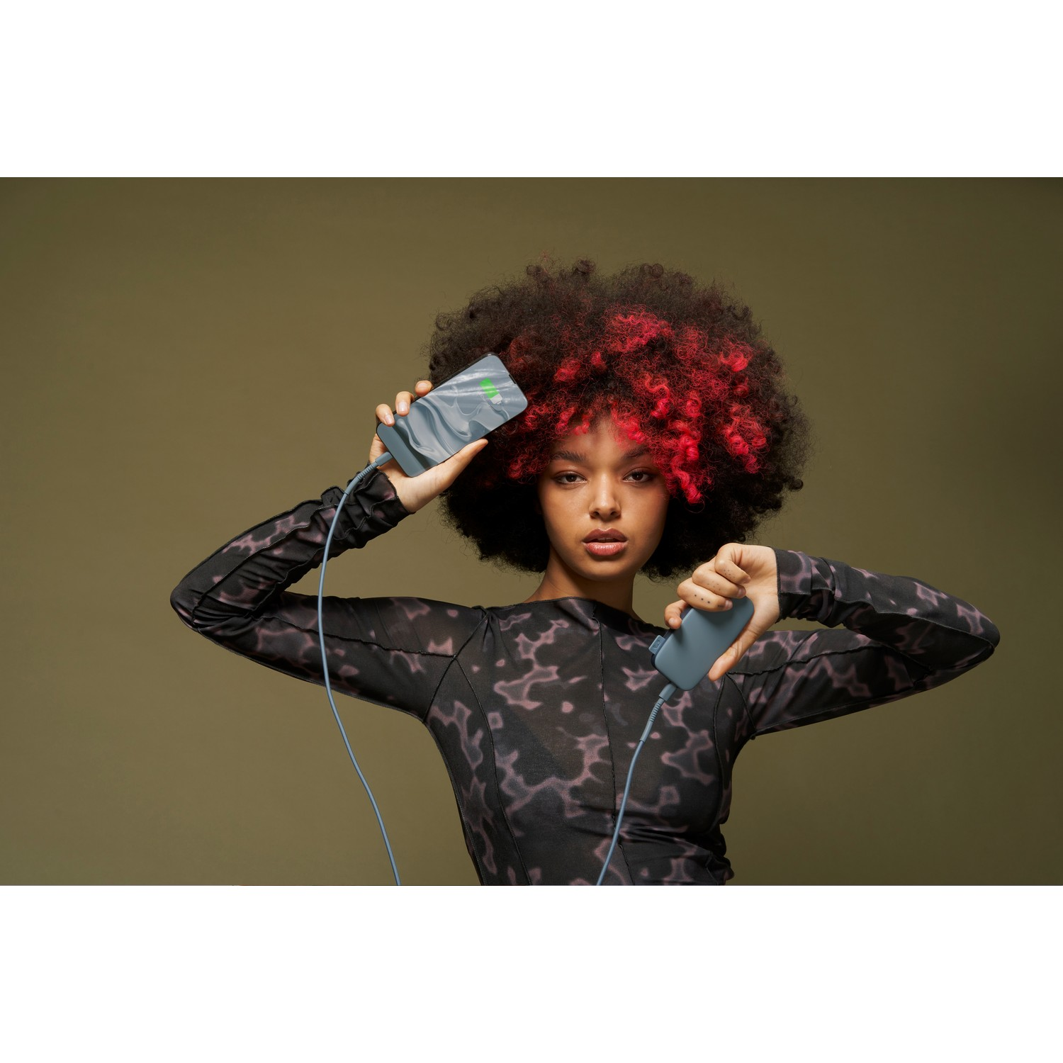 Femme tenant deux téléphones. L'un affiche une icône de batterie. Elle porte un haut à motifs et a les cheveux rouges.