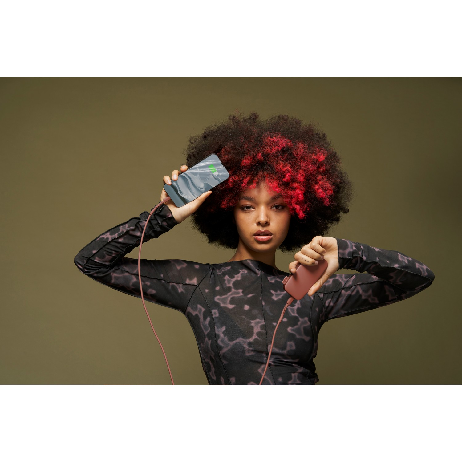 A woman holding a phone and a power bank. She has red highlights in her afro and wears a patterned top.