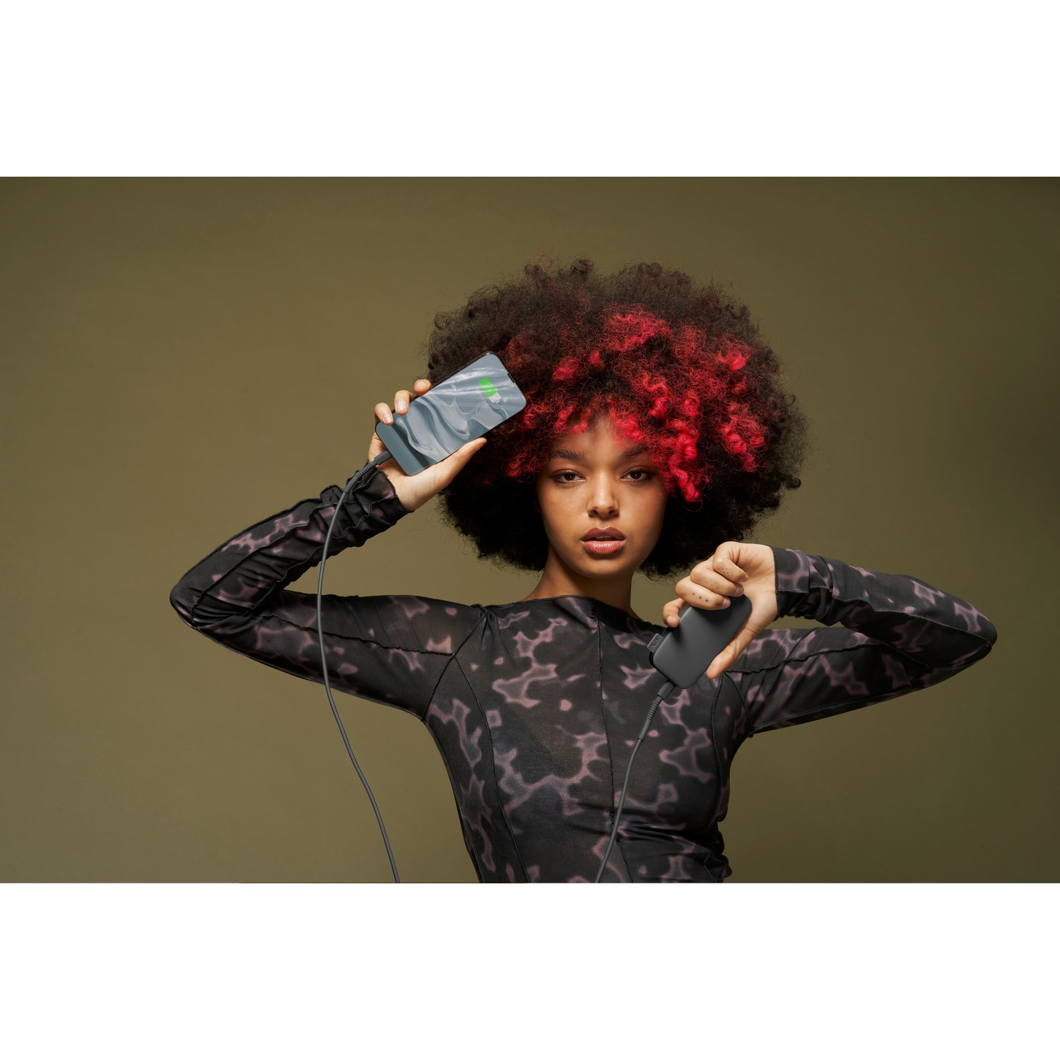 Een vrouw met rood gestreept haar houdt een oplaadtelefoon en een powerbank vast. Ze draagt een top met patroon op een groene achtergrond.