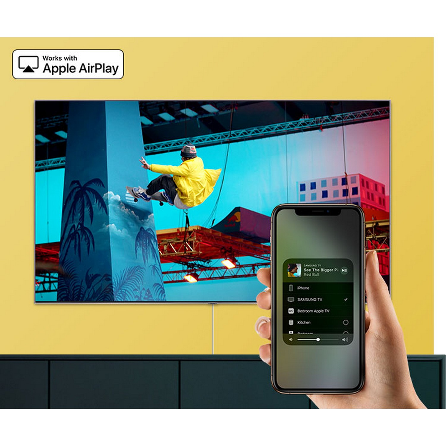 A person holding a phone showing Apple AirPlay controls, with a TV displaying a sports scene on a yellow wall.