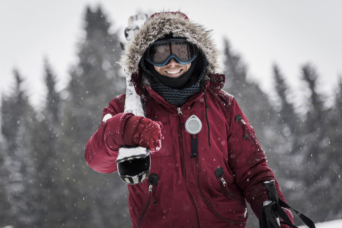 Mann in Winterausrüstung, der Skier hält. Schnee fällt, Wald im Hintergrund.