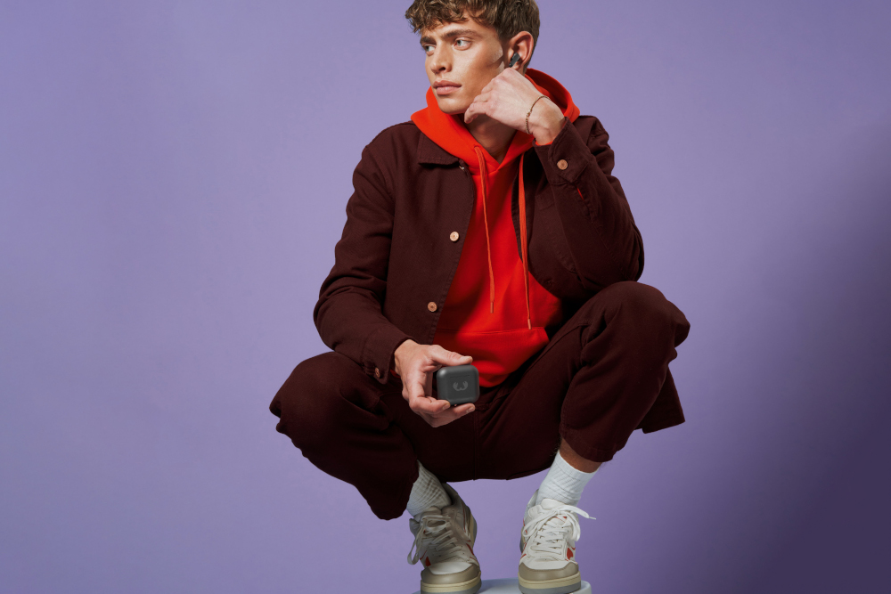 A man squats holding a gray earbud case, wearing red and brown clothes.