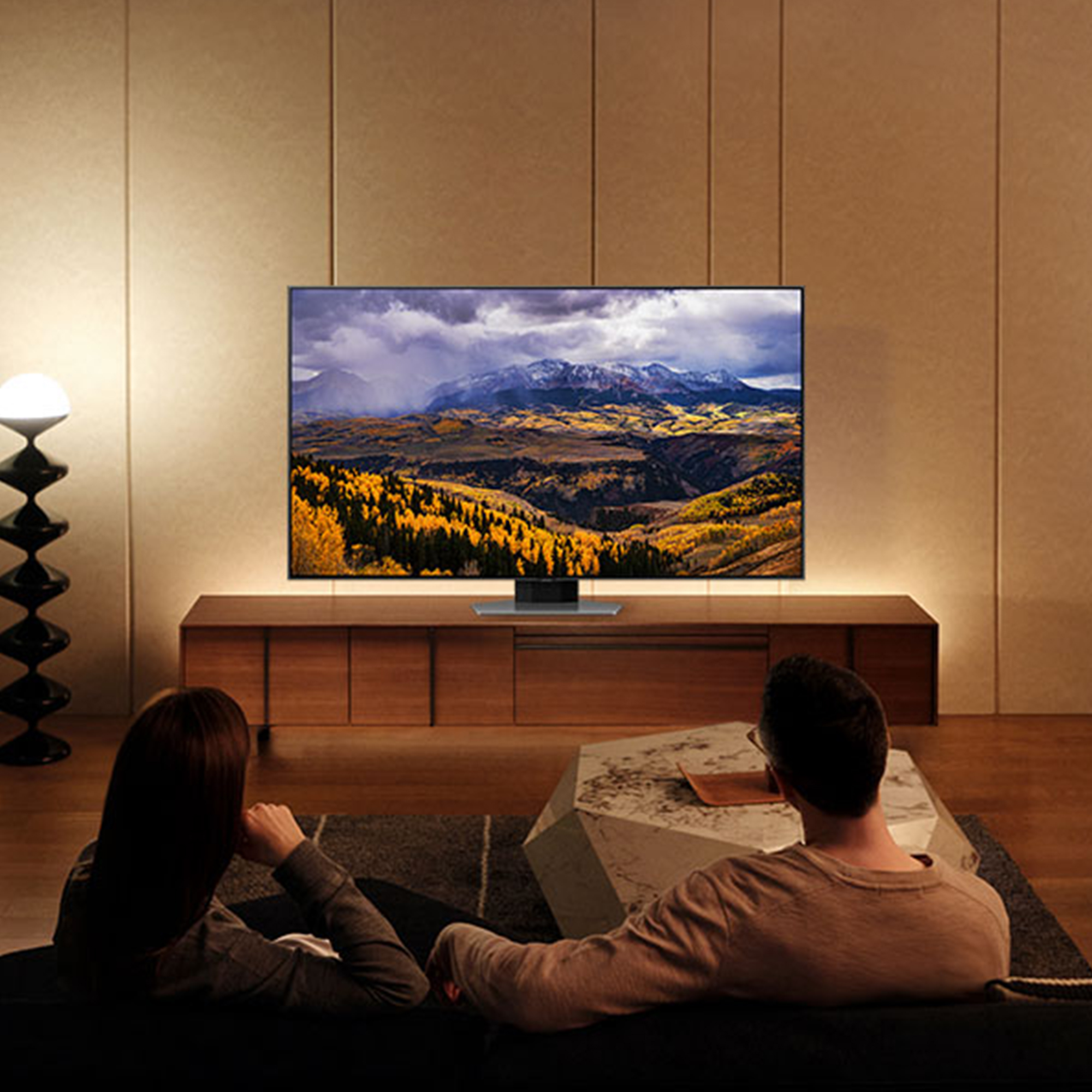 Dos personas viendo la televisión; paisaje de montaña en pantalla. Mueble de madera, sofá gris y decoración moderna.