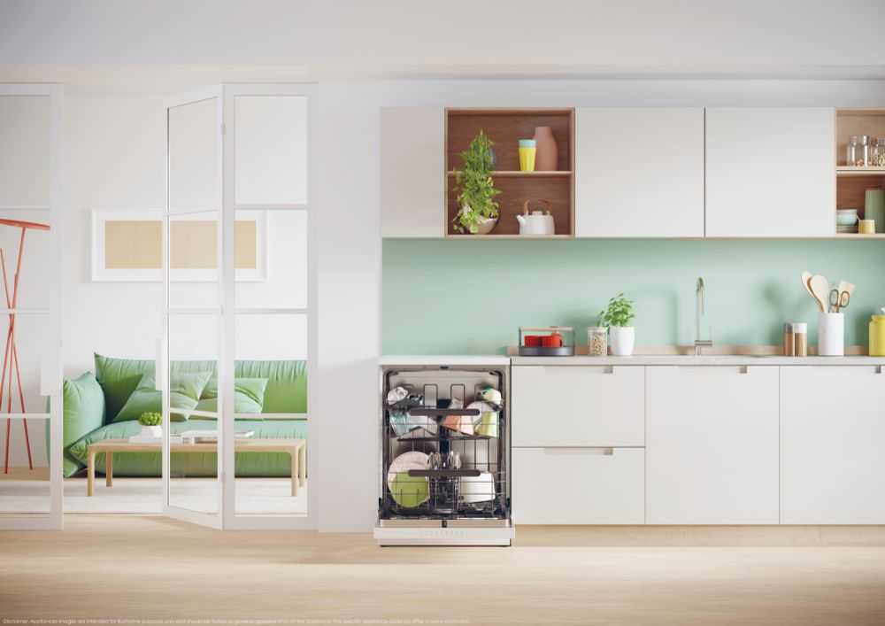 Interni cucina moderna. Lavastoviglie aperta, divano verde, mobili bianchi e pavimento in legno. Piante e piatti visibili.