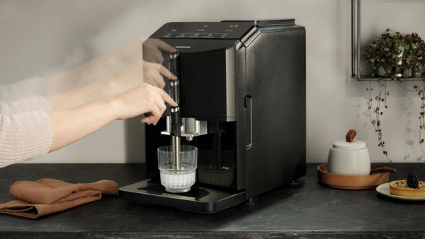 Eine Person bedient eine schwarze Kaffeemaschine. Ein Glas füllt sich mit Milch.