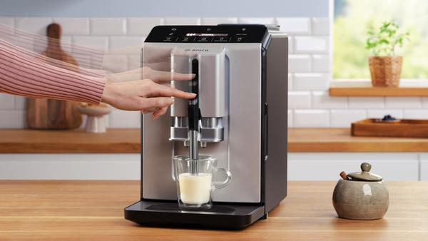 Eine Person bedient eine silberne Kaffeemaschine und gibt Milch in ein Glas ab. Eine Zuckerdose steht auf der Holztheke.