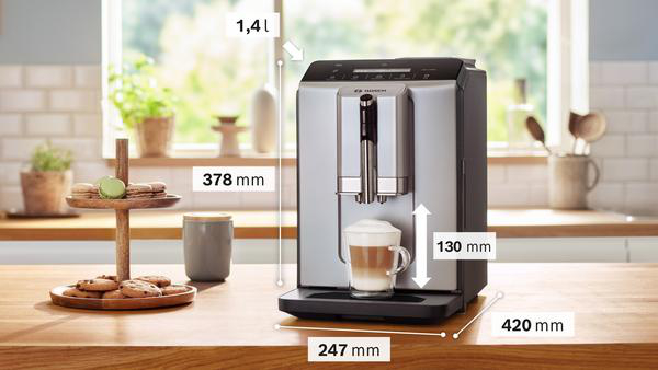 Eine silberne Kaffeemaschine mit einem Glas Latte. Kekse und Makronen auf einem Holzständer. Abmessungen sind mit Pfeilen angegeben.