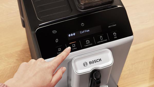 Eine Hand drückt die Kaffeetaste an einer schwarz-weißen Kaffeemaschine. Der Bildschirm zeigt 'Kaffee'.