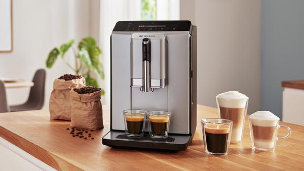 Eine Kaffeemaschine mit zwei Espressos. Kaffeebohnen in Papiertüten. Latte und Cappuccino in Gläsern auf Holzoberfläche.