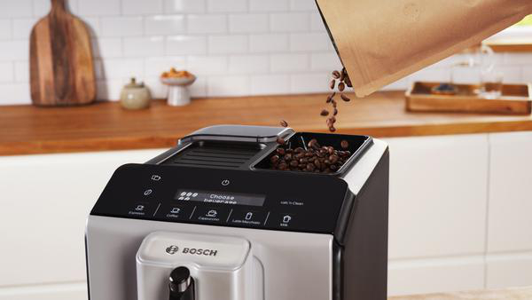 Kaffeebohnen werden in den Behälter einer Kaffeemaschine gegossen. Eine Tüte Bohnen wird gehalten. Küchenumgebung mit Holzplatte.