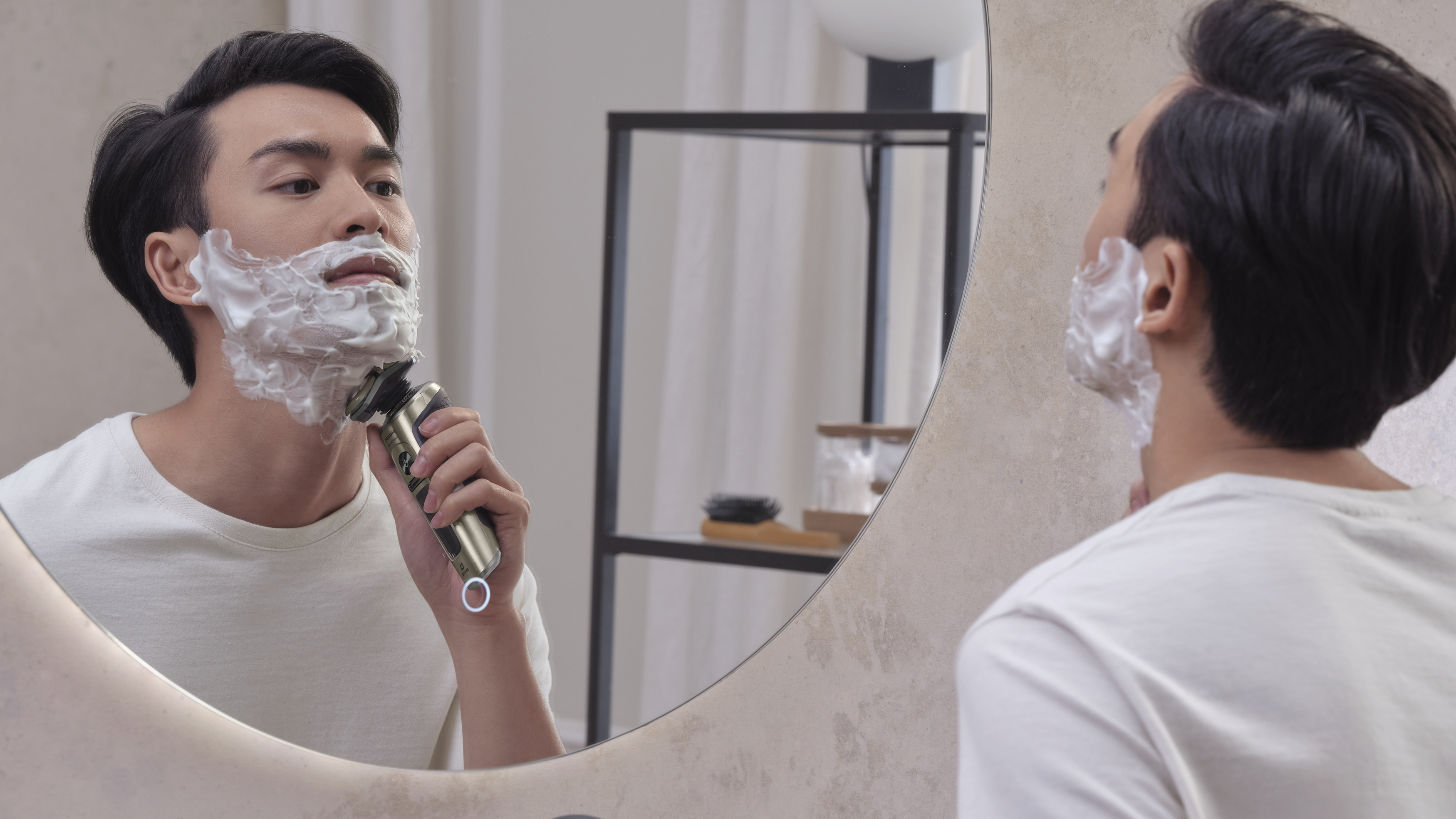 Mann rasiert sich im Spiegel, Schaum im Gesicht, Elektrorasierer in der Hand, Badezimmer-Szene, Profilansicht.