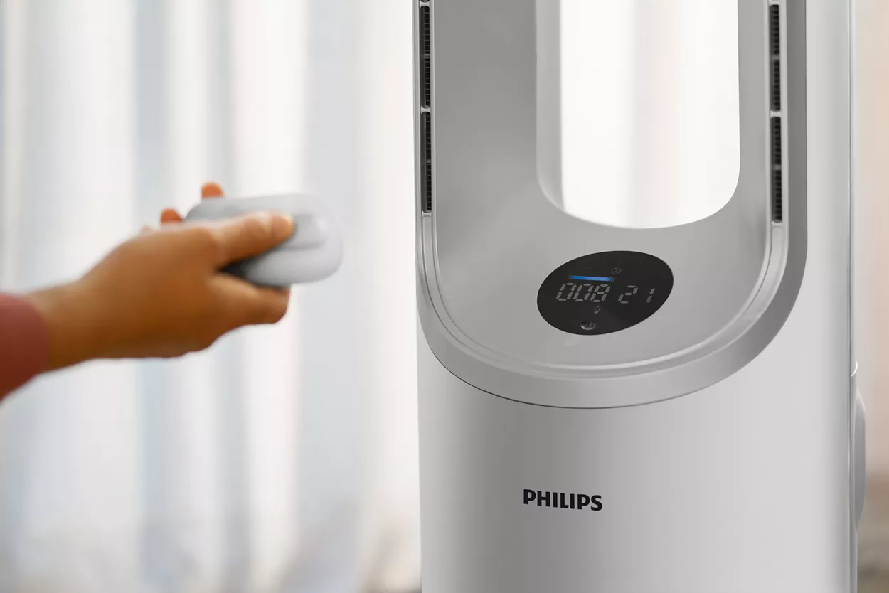 Egy személy keze tart egy távirányítót egy fehér Philips légtisztító közelében.