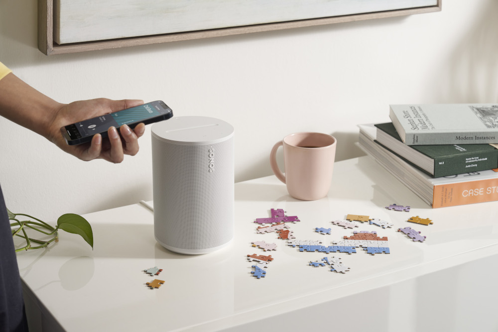 Persoon gebruikt telefoon naast een speaker, puzzel en roze mok.