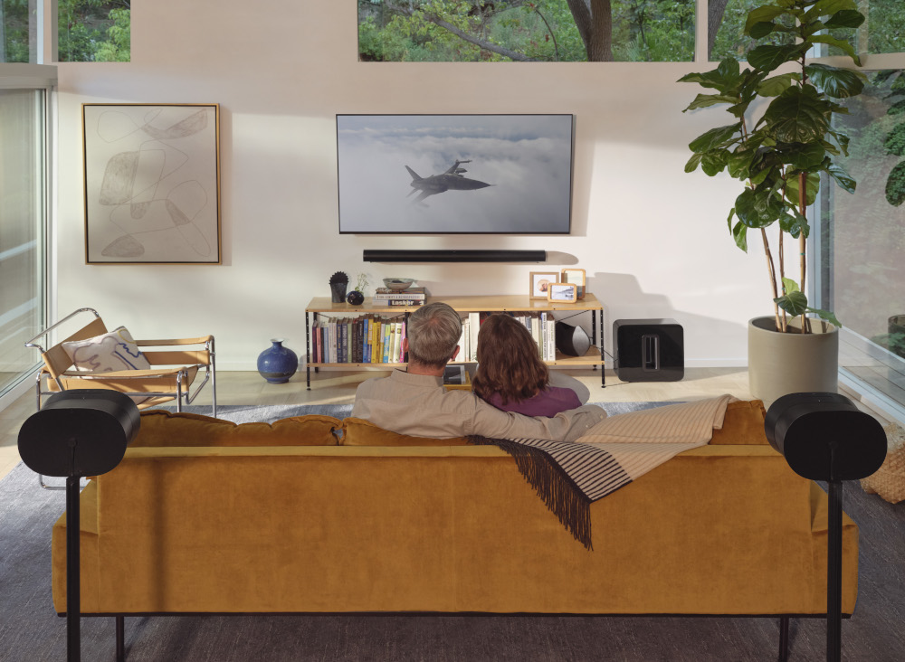 Man and child watch TV. The room has a couch, speakers, and plants.
