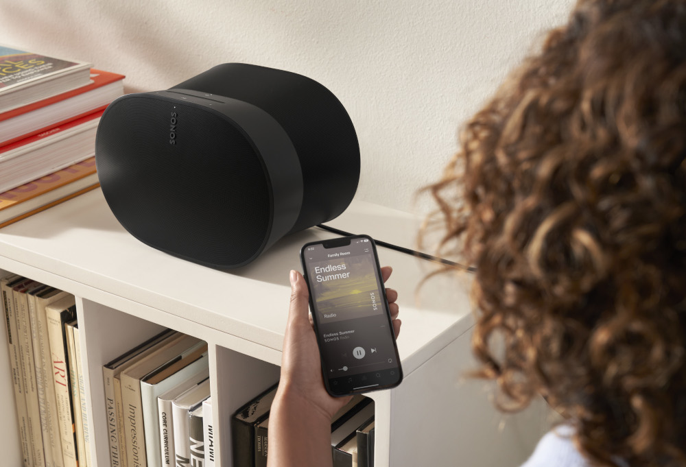 Black speakers and phone playing music. Person holding phone. Bookshelf in the background.