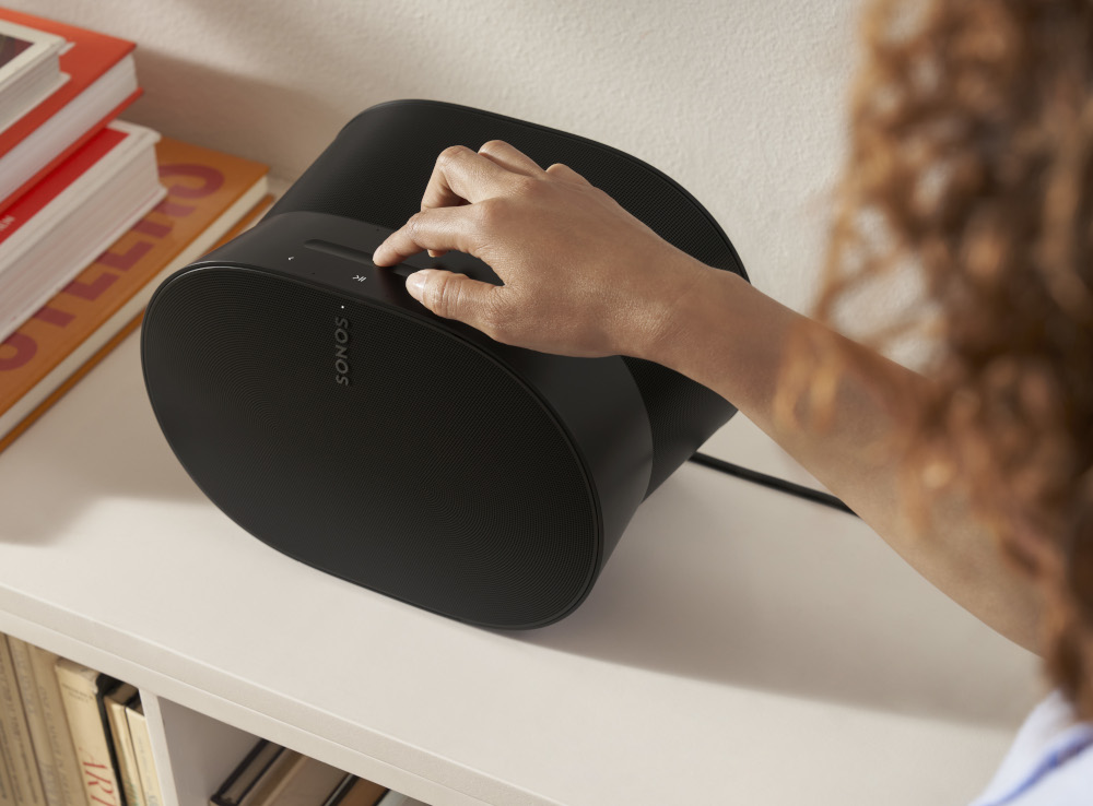 Person's hand adjusting volume on a black Sonos speaker on a shelf with books.
