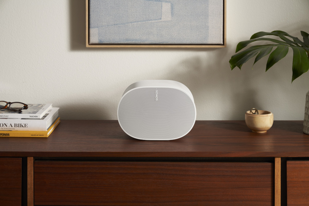 A white Sonos speaker on a wooden surface, with a painting and plant behind.