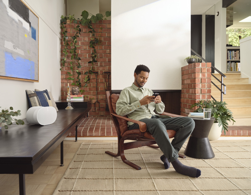 Man sitting in a chair looking at his phone. In a living room setting. Bookshelf, fireplace.
