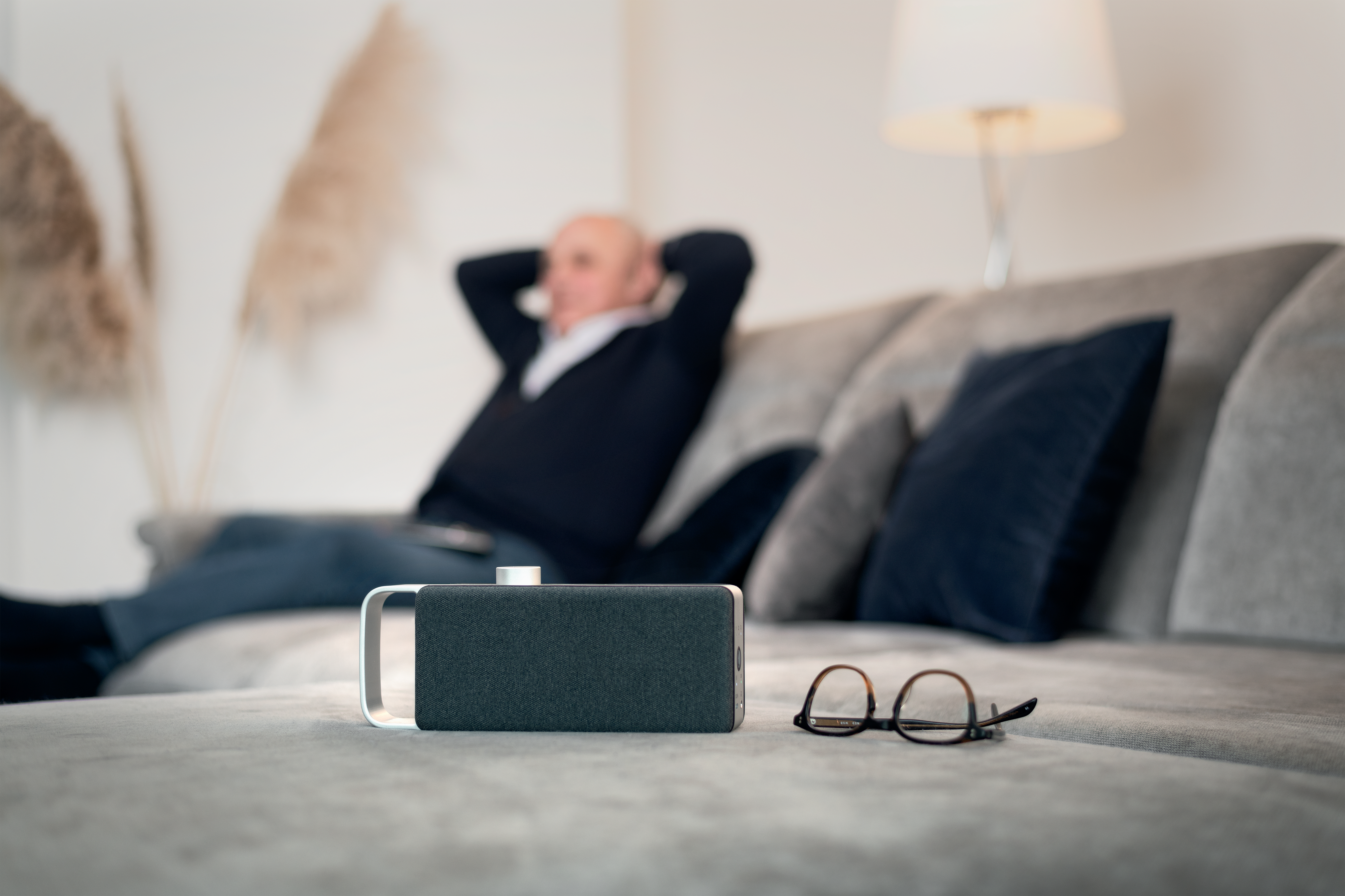 Ein Mann entspannt sich auf einem grauen Sofa, mit einem Lautsprecher und einer Brille auf dem Sofa. Verschwommener Hintergrund.