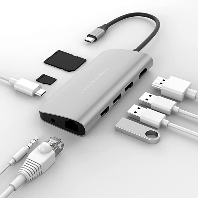 Concentrador USB-C plateado con múltiples puertos y cables conectados, junto a tarjetas SD y una unidad USB.