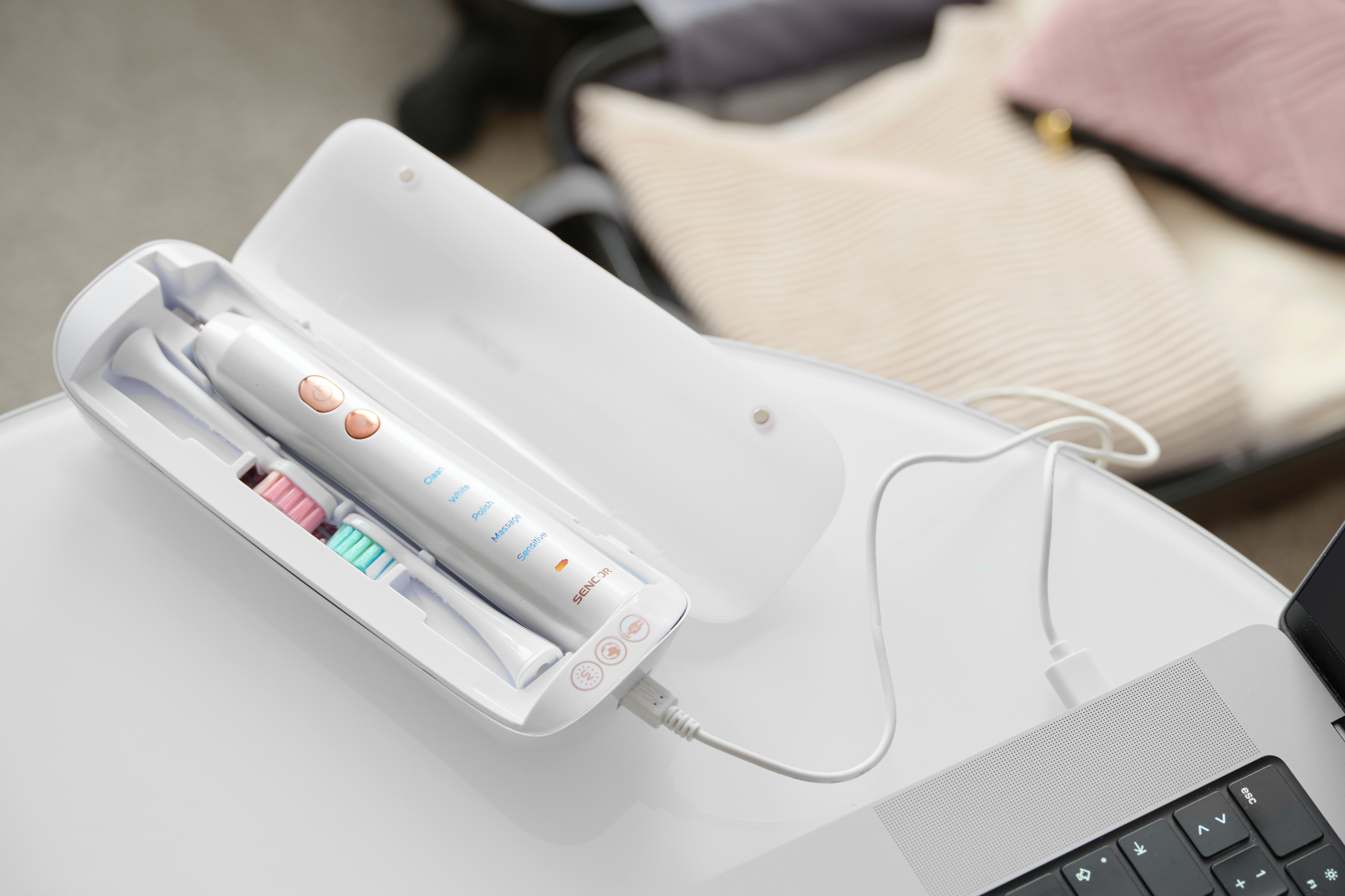 Fehér elektromos fogkefe nyitott tokban, töltőkábellel egy fehér felületen, laptop mellett.
