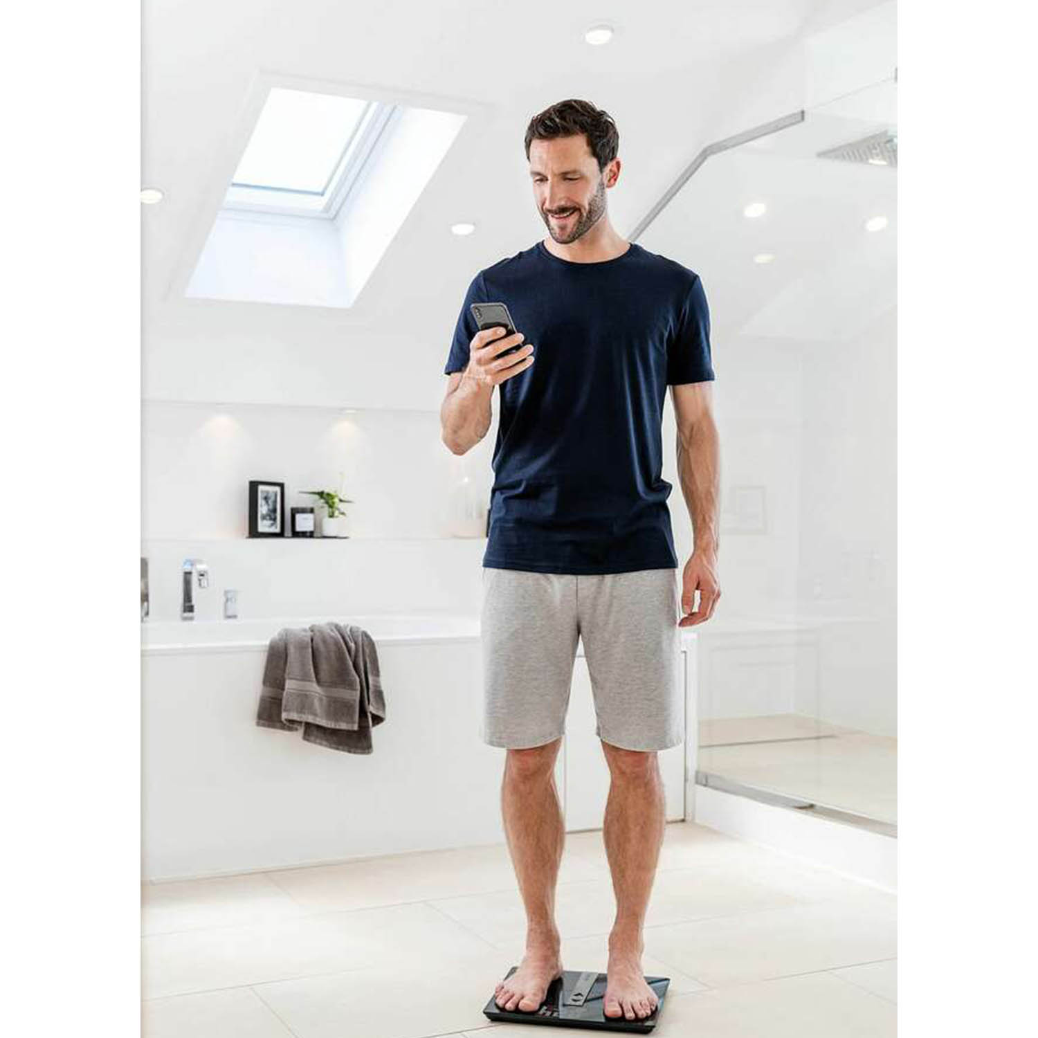 Uomo in piedi su una bilancia intelligente, che guarda il suo telefono in un bagno.