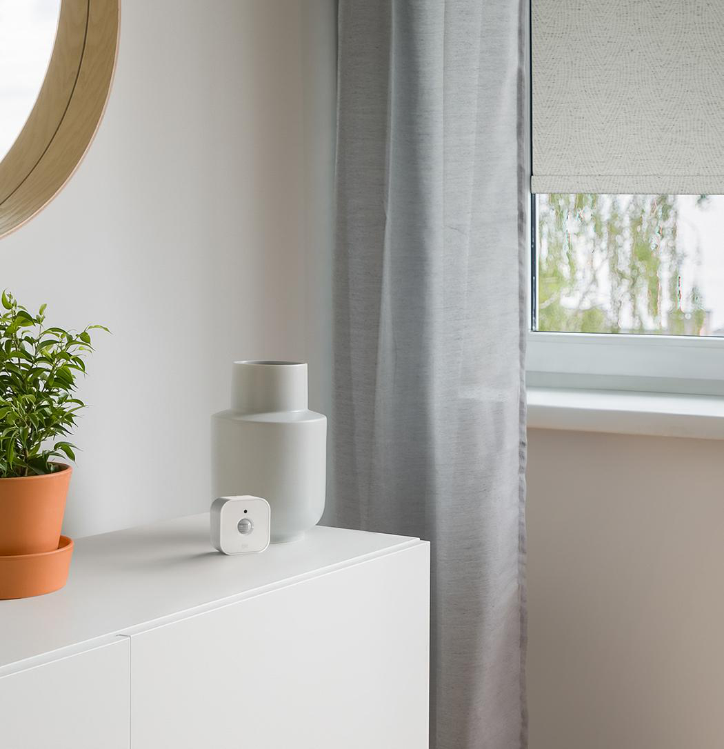 Ein weißes Regal mit einer Pflanze im Topf, einer weißen Vase und einem kleinen quadratischen Sensor vor einer Wand.